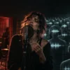 A human vocalist stands apart from rows of identical synthetic listeners in a recording studio